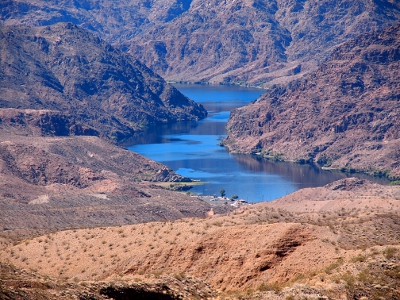 [Ribbon of water passing through the lowest portion of a winding canyon comprised of medium dark rock. A parking lot near the lake is a spec in the photo.]